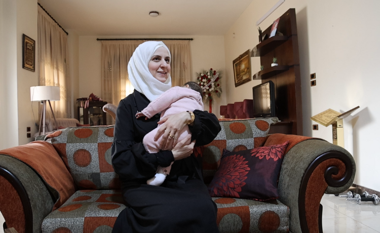 A woman in a white headscarf holds a newborn wearing a pink outfit. They are in a living room.